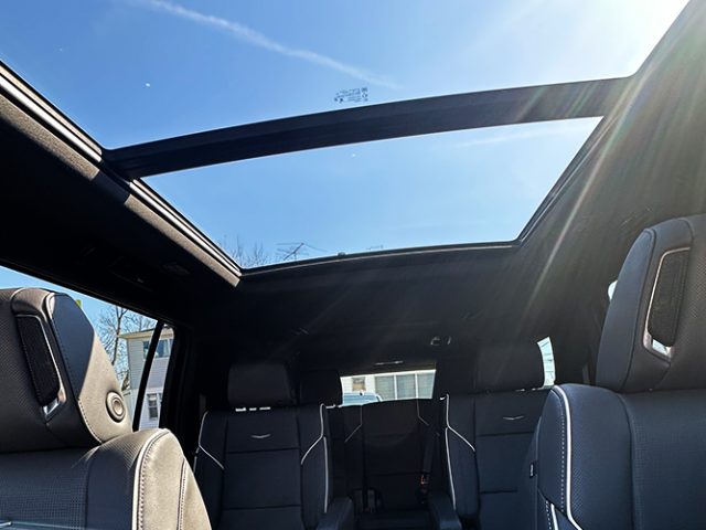 Inside a car with a large panoramic sunroof open, showing blue sky and sunlight streaming in toward the rear seats.
